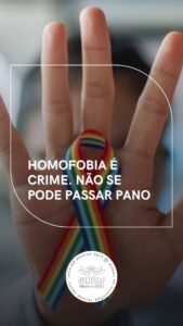 Imagem de um mão com o símbolo da fitinha da homofobia. No meio temos o título Homofobia é crime. Não se pode passar pano. Para fechar a peça a logomarca do SINPAF. 