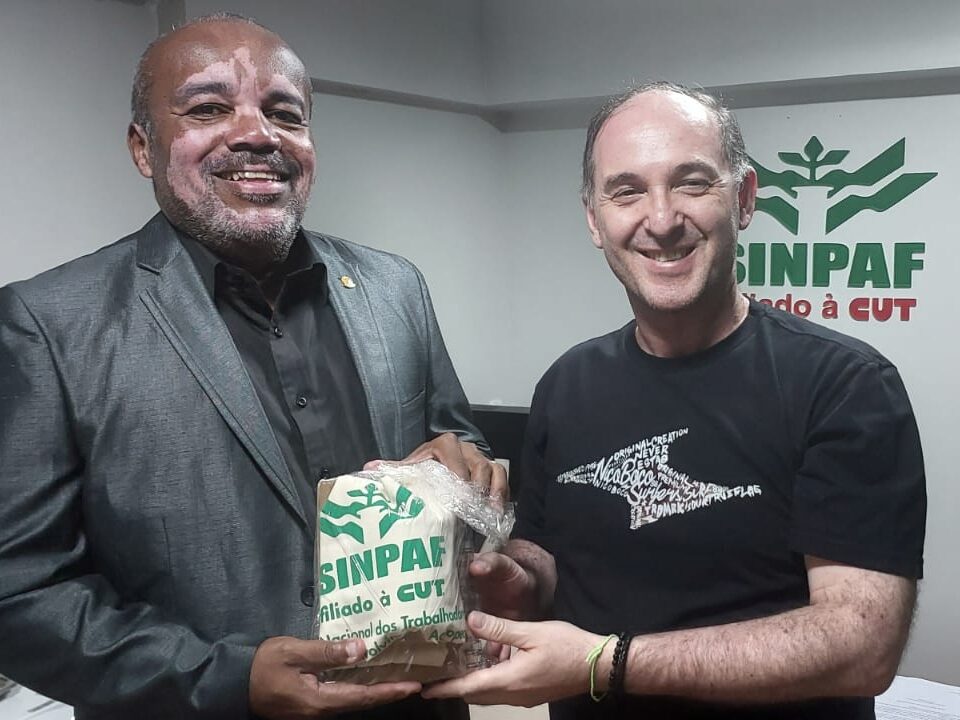 Foto do presidente do SINPAF Marcus Vinicius. Ele é um homem branco e calvo. Ao lado dele o diretor de saúde do SNTPV. Rogério Amaral. Rogério Amaral é um homem negro e tem algumas manchas brancas no rosto. Ele é um homem calvo. O presidente do SINPAF entrega a lembrança do sindicato para o diretor do SNTPV. 