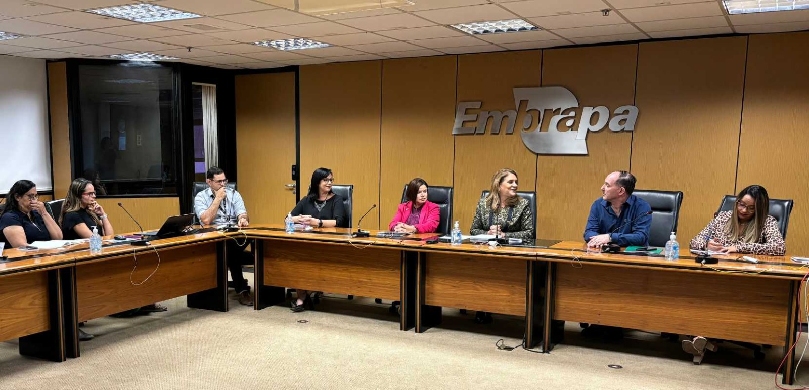 Reunião na Embrapa com a diretoria executiva. As pessoas estão na mesa e temos o símbolo da Embrapa em prateado em uma parede de madeira palha.