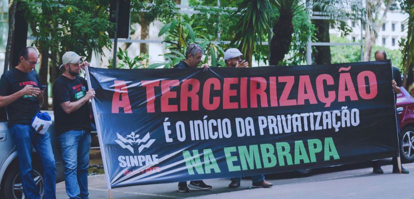 Faixa no meio da cor preta e escrito A Terceirização é o início da privatização na Embrapa. Dois homens do lado esquerdo e um do lado direito segurando a faixa.