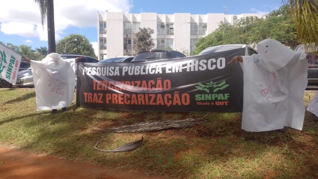 Dois fantasmas (homens vestidos de branco inclusive a cabeça) seguram a faixa contra a tercerização. Está escrito Pesquisa Pública em Risco. Terceirização traz precarização. 