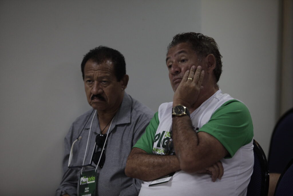 Dois homens pardos. Eles estão sentados. Um deles com camisa cinza e com bigode. O outro com a mão esquerda no rosto. Eles estão na Plenária Regional do Nordeste do SINPAF