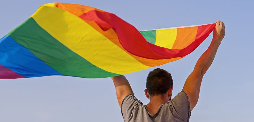 homem de costas com os dois braços levantados segurando a bandeira LGBTQIAPN+